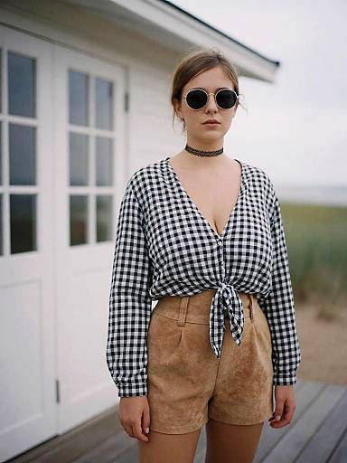 Photograph of a young woman with fair skin, brown hair in a bun, wearing black-and-white checkered blouse, tan shorts, black choker