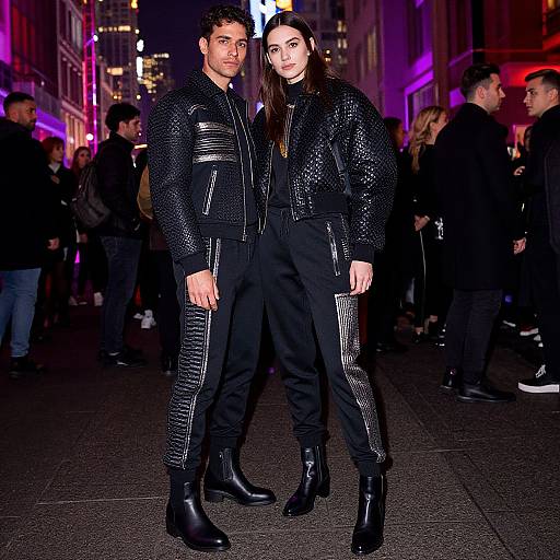 Photograph of a tall, dark-haired couple in black leather jackets and pants, standing in a neon-lit urban street at night, surrounded by other