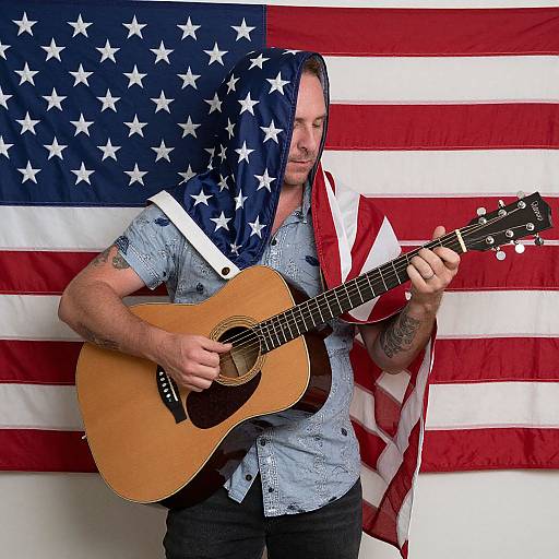 Photograph of a tattooed man playing an acoustic guitar, wearing a blue shirt with a draped American flag scarf and Stars and Stripes background.