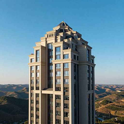 Photograph of a tall, beige, modern high-rise building with reflective windows, set against a clear blue sky and rolling hills.