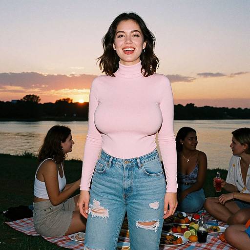 Photograph of a smiling woman with dark hair in a pink long-sleeve top and ripped jeans, standing at sunset by a lake, surrounded by