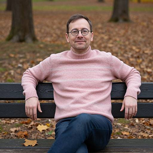 Photograph of a middle-aged man with glasses, short brown hair, and light beard, wearing a pink sweater and blue jeans, sitting on a black
