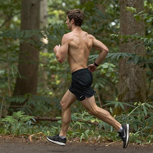 Photograph of a muscular, shirtless man jogging in a lush, green forest. He wears black shorts, black sneakers, and a watch, with