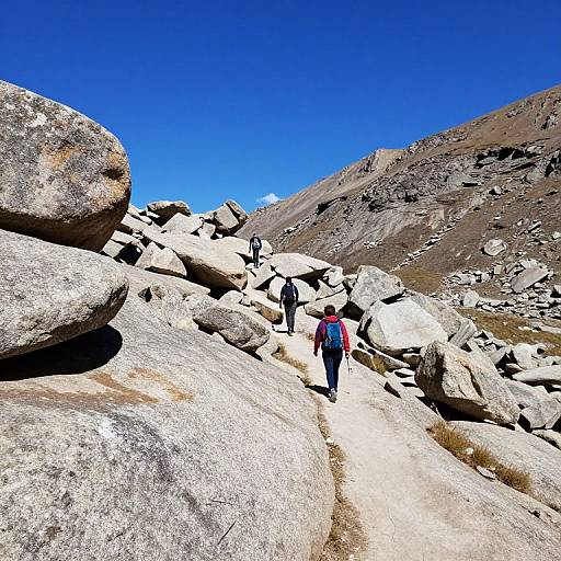 Photograph of two hikers in mountainous terrain with large, sunlit gray boulders, wearing backpacks and hiking gear, under a vivid