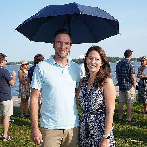 Photograph of a smiling couple standing outdoors under a black umbrella; man in light blue polo, woman in patterned dress, people and lake in background