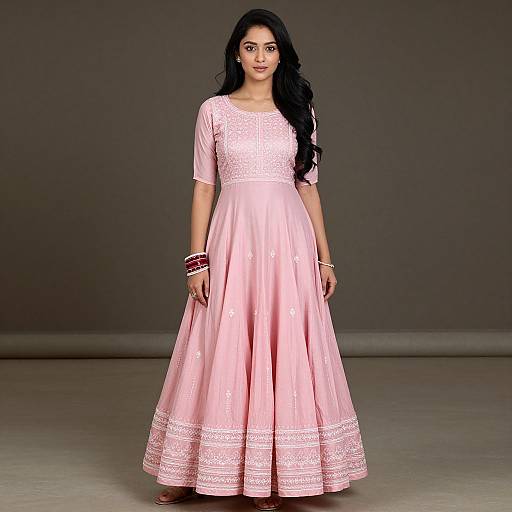 Photograph of a young woman with long black hair wearing a pink, knee-length, lace-embellished dress, standing against a gray background.