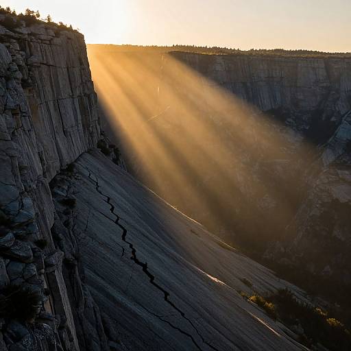 Photograph of a dramatic sunset over a deep canyon, with golden sunlight streaming through the cliffs, casting long shadows on the rugged, textured rock face.