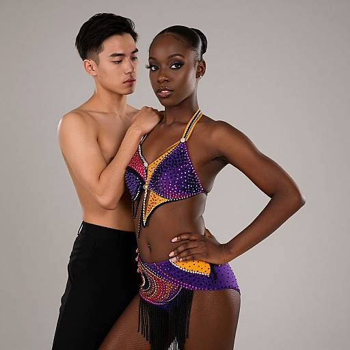 Photograph of a shirtless Asian male dancer with short black hair, standing behind a dark-skinned female dancer in a colorful, beaded, fringe