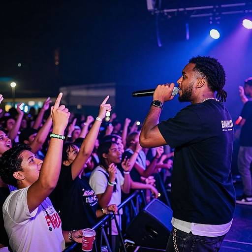 Photograph of a male rapper with dreadlocks performing on stage, crowd raising hands, vibrant blue and purple stage lights, wearing black shirt, white wrist