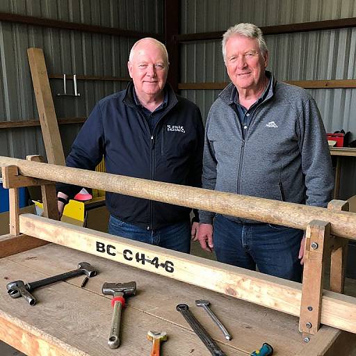 Photograph of two older Caucasian men in a wooden workshop, standing beside a workbench with tools and a labeled beam.