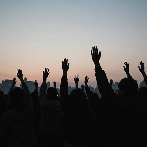 Silhouetted crowd with raised hands at sunset, city skyline in the background. Photograph captures collective energy and anticipation.