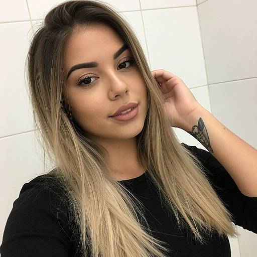 Photograph of a young woman with long, ombre blonde and brown hair, wearing a black top, smiling softly, with a subtle tattoo on