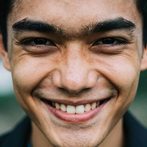 Close-up photograph of a smiling young man with dark eyebrows, brown skin, and white teeth, wearing a black shirt, against a blurred background.