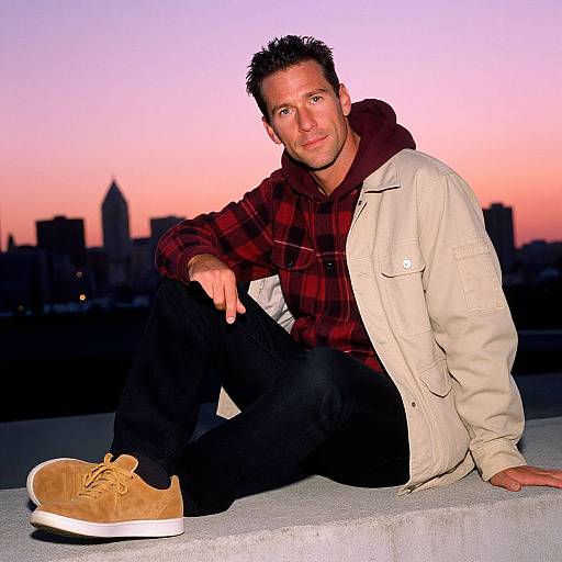 Photograph of a smiling man with short dark hair, wearing a red plaid shirt, beige jacket, and tan sneakers, sitting on a rooftop at
