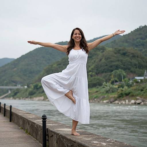 Photograph of a smiling woman with long dark hair, wearing a white sleeveless dress, balancing on one foot with arms outstretched, standing on