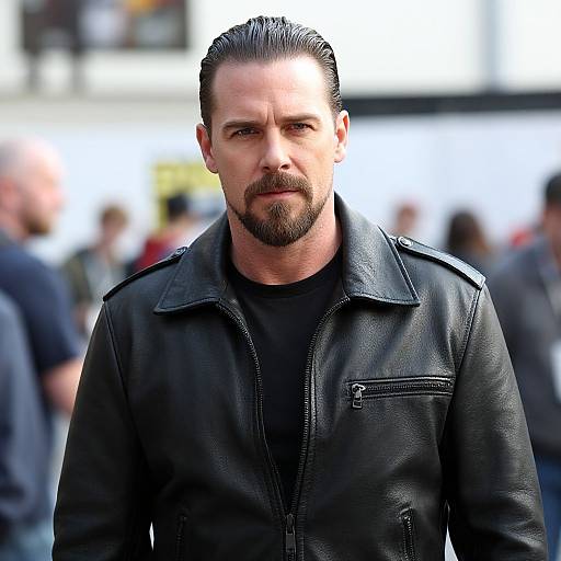 Photograph of a serious-looking, bearded man with slicked-back hair, wearing a black leather jacket, standing in a blurred, bustling outdoor crowd