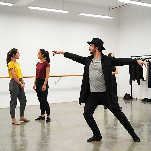 Photograph of a modern dance class: a bearded man in a black outfit and cap gestures to two women in yellow and red shirts, standing in