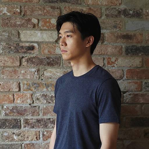 Photograph of an Asian man with short black hair, wearing a plain black t-shirt, standing against a rustic, multi-colored brick wall, looking to