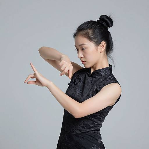 Photograph of an Asian woman with black hair in a bun, wearing a black lace sleeveless dress, gracefully pointing and stretching against a plain light blue