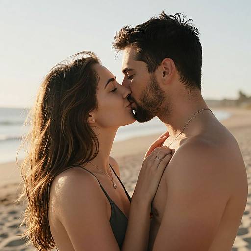 Photograph of a shirtless man and woman kissing on a sunlit beach, both with long brown hair, wearing necklaces, and standing close with