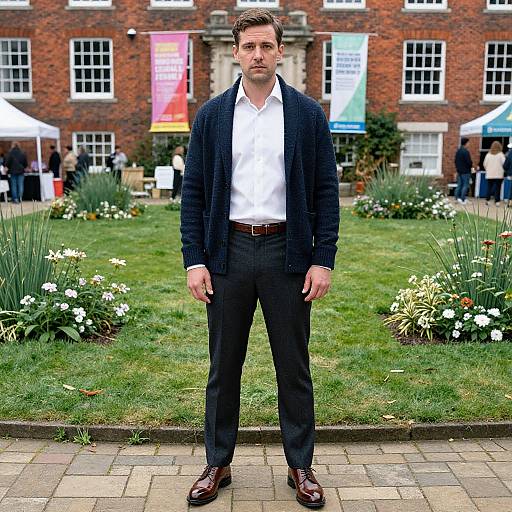 Photograph of a tall, handsome man with short brown hair, wearing a black cardigan, white shirt, black pants, and brown shoes, standing