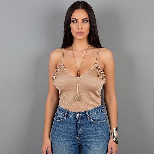 Photograph of a brunette woman with long hair, wearing a beige tank top with fringe, blue jeans, and a long necklace, standing against a plain