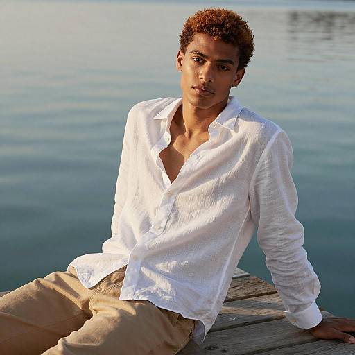 Photograph of a young man with short curly brown hair, light brown skin, wearing a white button-up shirt and beige pants, sitting on a wooden