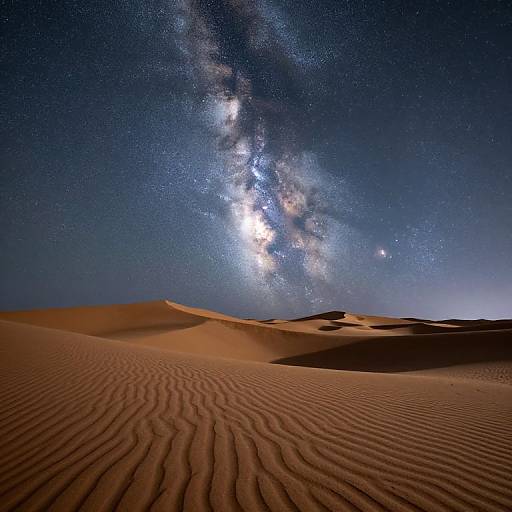 Photograph of a desert night sky with rippled sand dunes, Milky Way galaxy visible, bright stars, and a clear deep blue sky.