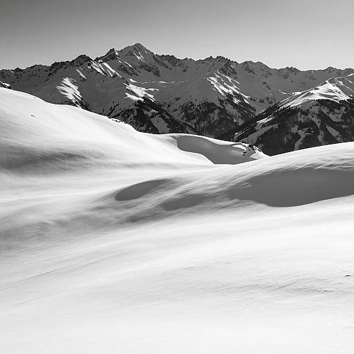 Black-and-white photograph of a snowy mountain range with sharp, jagged peaks in the background and smooth, white snowdrifts in the foreground.