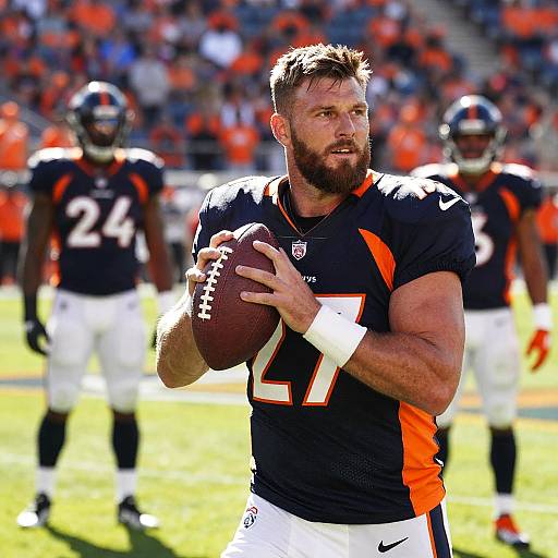 Photograph of a muscular, bearded male football player in black and orange uniform holding a football, with blurred teammates and a crowd in the background on
