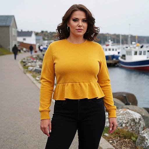 Photograph of a confident woman with wavy brown hair, wearing a bright yellow long-sleeve top and black pants, standing on a waterfront path