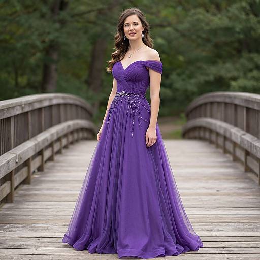 Photograph of a smiling woman with long brown hair in an off-shoulder, purple, full-length, tulle gown standing on a wooden bridge