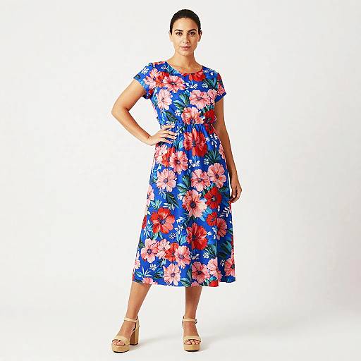 Photograph of a woman with dark hair in a blue dress with large red, pink, and white floral patterns, standing confidently, hand on hip,