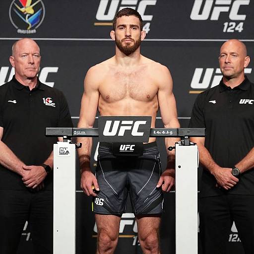 Photograph of a muscular, shirtless male UFC fighter with dark hair and beard, wearing black shorts, standing between two officials, UFC logo and weight