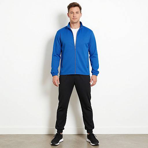 Photograph of a young man with short brown hair, wearing a blue zip-up jacket, white shirt, black pants, and black sneakers, standing against