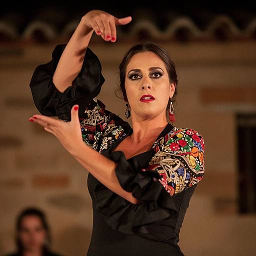 Photograph of a dark-haired woman with red lipstick, wearing a black dress with colorful embroidered shoulders, mid-dance, arms raised, red nails,