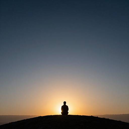 Silhouetted person sitting on hilltop at sunset, backlit with a bright orange sun against a deep blue sky. Minimalistic photograph.