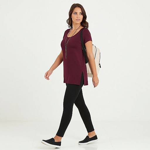 Photograph of a brown-haired woman with medium skin tone, wearing a maroon top, black pants, black flats, and a white backpack, walking