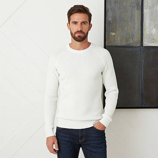 Photograph of a handsome, bearded man with short brown hair, wearing a white ribbed sweater and dark jeans, standing against a white wall with