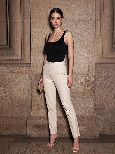 Photograph of a confident woman with fair skin and dark hair, wearing a black tank top, white high-waisted pants, and nude heels,