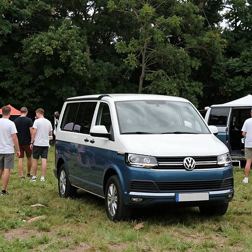 Photograph of a blue and white Volkswagen van parked on grass, with several casually dressed people standing nearby, surrounded by trees.