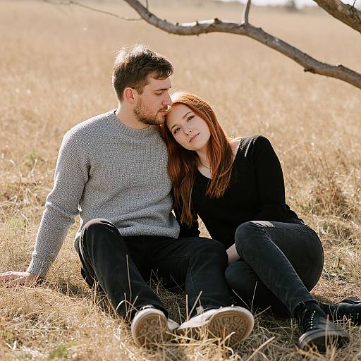 Photograph of a young couple sitting in a sunlit, dry grass field; man in gray sweater, woman in black, she leans on him affection