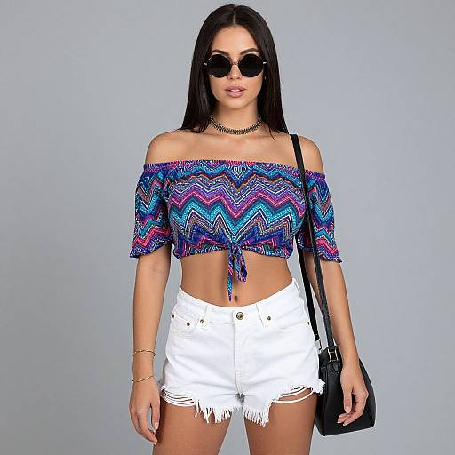 Photograph of a young woman with long black hair, wearing round sunglasses, a colorful off-shoulder crop top, white frayed shorts, black