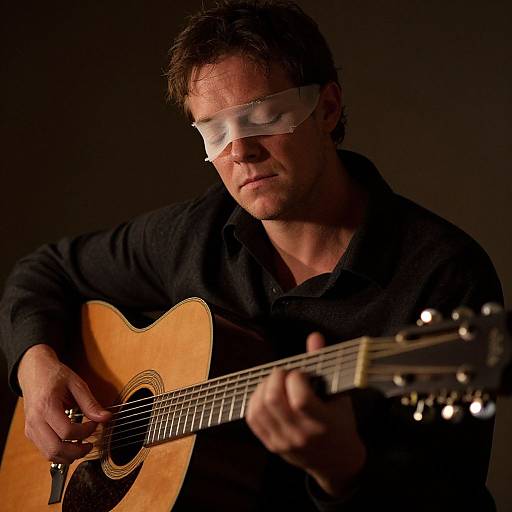 Photograph of a man with short brown hair, wearing white sunglasses and a black polo shirt, intensely playing a wooden acoustic guitar in low, dramatic lighting