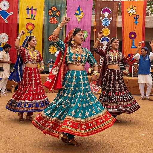Photograph of three Indian women in vibrant traditional lehengas, gold jewelry, and red-orange headscarves, dancing energetically in a colorful