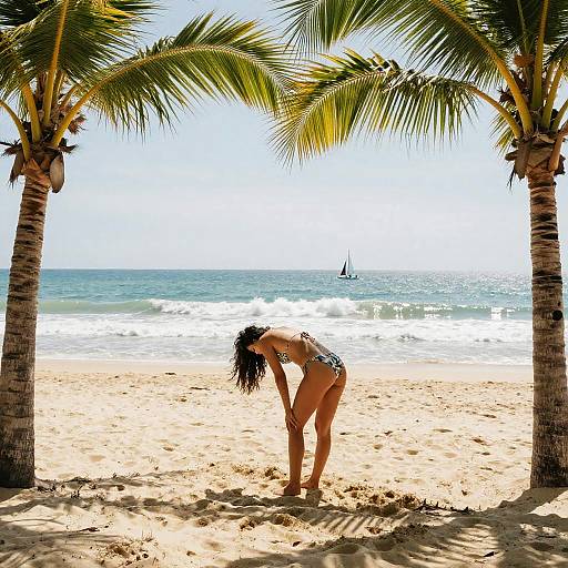 Photograph of a woman in a black and white bikini, bending over on a sunny beach, framed by two palm trees, with a sailboat on
