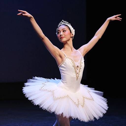 Photograph of an elegant ballerina in a white tutu with gold accents, tiara, and poised arms, performing on a dark stage.