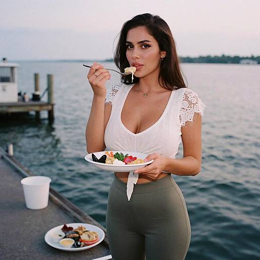 Photograph of a fit woman with dark hair, wearing a white lace crop top and gray pants, eating salad by a lakeside.
