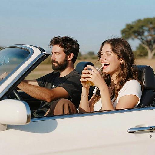 Photograph of a smiling brunette woman in a white t-shirt, holding a drink, seated in a white convertible, driven by a bearded man in