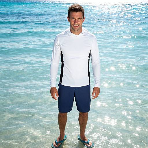 Photograph of a smiling man with short brown hair, wearing a white long-sleeve shirt, black shorts, and teal flip-flops, standing